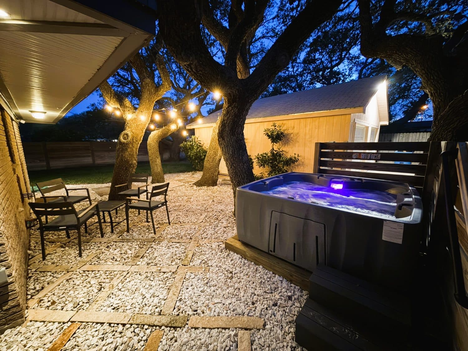 Finished hot tub area at dusk: stone and gravel patio, string lights, seating, and the tub lit and running